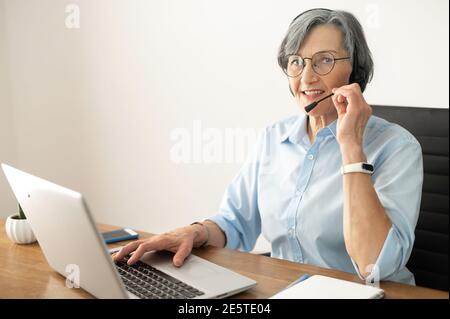 Senior mature female gray-haired call center operator sitting at the desk in the home office, wearing a headset, answering the call, listening to the Stock Photo