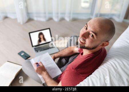 A bald young happy man is working at a laptop and writing something down on paper. Top view. The concept of online courses and telecommunication. Stock Photo