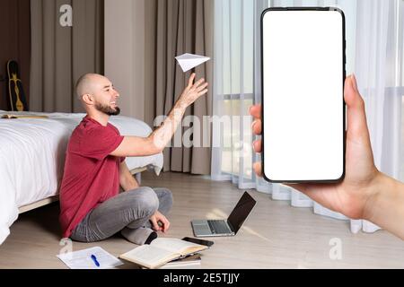 A young smiling man sits on the floor and launches a paper airplane into the air. On the floor is a laptop, books and papers. Smartphone with mock up. Stock Photo