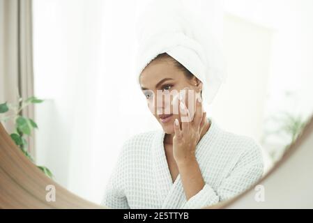 Beautiful young woman applying cream in bathroom. Face cream. Skin care. Beautiful smiling woman with a towel on her head after showering. Stock Photo