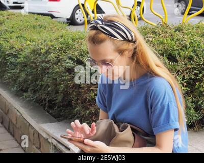 Redhead girl playing games on mobile phone while sitting on city stree Stock Photo
