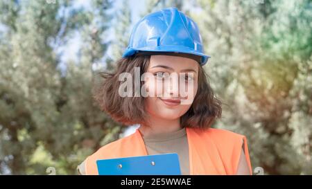 Portrait of a cheerful young woman engineer or worker in a uniform and protective blue hardhat, smiling and looking at camera at construction site Stock Photo