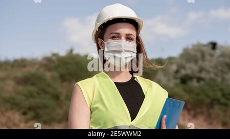 Portrait of young female construction worker or engineer with white helmet and face mask looking at camera at construction site. Stock Photo