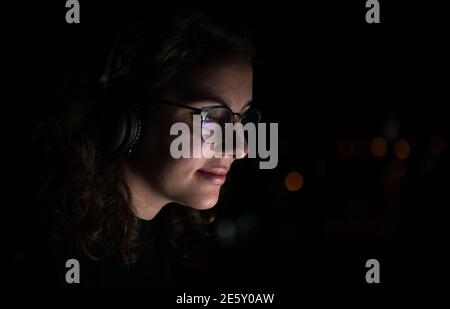 Woman with glasses and wireless headphones surfing the internet while it is night, the computer screen is reflected in her glasses. Stock Photo