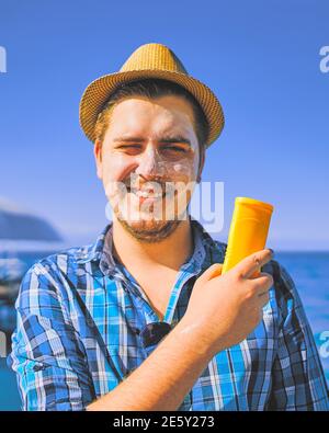 Guy putting on sun protection lotion on face. Man tanning using sun block body cream on summer beach. Care for sensitive skin during vacation. Funny man in straw hat and shirt on sea background. Stock Photo