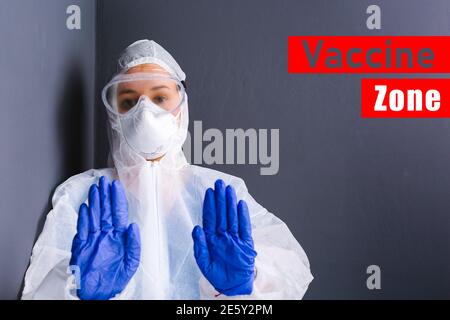 A young doctor in protective equipment and medical goggles shows stop with both hands against the background of a gray wall and the inscription Stock Photo