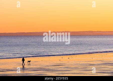 A single person walks their dog on an empty beach as the sun sets, the warm colours of the sky reflected in the wet sands.. Stock Photo