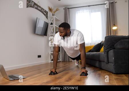 African American guy holding his body in plank position, using push up bar to do push-ups on the floor in the living room while isolated during Stock Photo