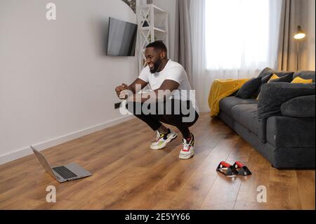 Handsome young African American is holding his body in the squat position, getting active while isolated, learning from online yoga course, fitness Stock Photo