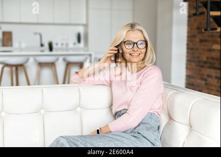 Stylish smart middle-aged woman wearing eyeglasses has phone conversation, a business lady relaxed sits on the couch at modern apartment, laugh and Stock Photo