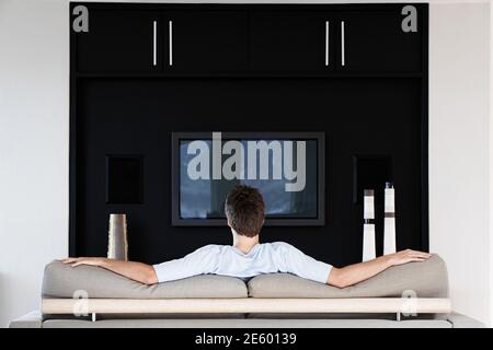 Rear view of young man watching television while sitting on couch in living room Stock Photo