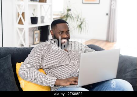 Portrait of smiling young African American guy placed the laptop on knees while relaxing on the sofa, typing email, messaging friend, freelancer Stock Photo