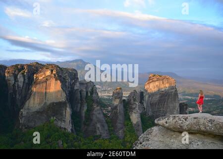Landscape with a blonde young girl on a red dress overlooking the Meteora Holy Rocks in Thessaly, Greece. Stock Photo
