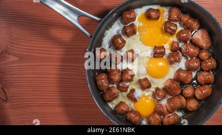 Banner with fried sausages with chicken eggs in process of frying in a frying pan at the red wood table with copy space for text Stock Photo