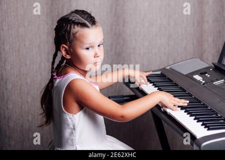 a pensive, dreamy girl in a white summer dress and two pigtails trains chords on an electronic synthesizer in a home interior Stock Photo