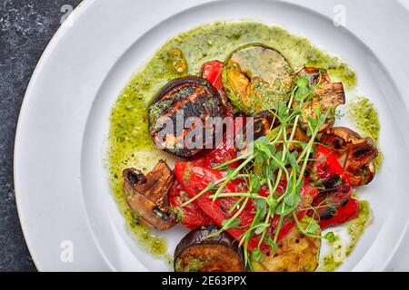 Grilled baked vegetables, eggplant, zucchini, bell pepper, champignon mushrooms, on a white plate against a dark background, with microgreen Stock Photo