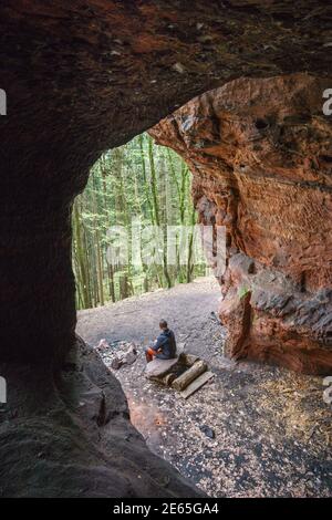 young man sitting on bank in front of red sandstone cave with view into the forest near Trier, Germany Stock Photo