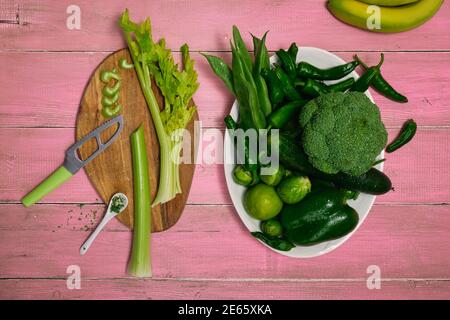 Vegetables such as celery, broccoli, peppers and cucumbers in a white dish on a pink wooden surface. On a cutting board the knife cut the celery Stock Photo
