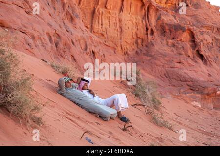 Wadi Rum Desert, Jordan 03-31-2010: Two young Arabic men wearing traditional dress are lying on the sandy slope on the foothills of red sandstone clif Stock Photo