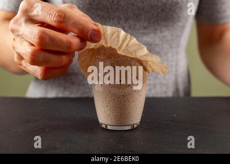 A woman is carefully removing the top cover of a sourdough starter culture which is about to overflow the glass cup. The gooey starter culture sticks Stock Photo
