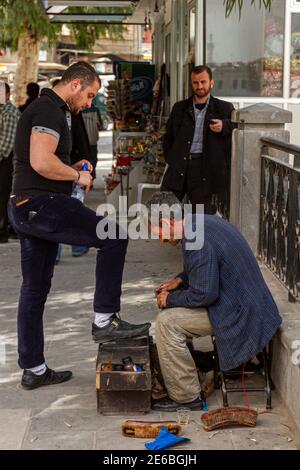 Hama, Syria 04-02-2010: An elderly Syrian Shoe shiner is polishing a young man's shoes on his shoeshine box on the sidewalk by a crowded street. Old m Stock Photo