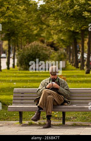 Haarlem, Netherlands 06-16-2010: A young stylish man wearing vintage clothes and a flat cap hat is sitting crosslegged alone on a park bench. He is sm Stock Photo
