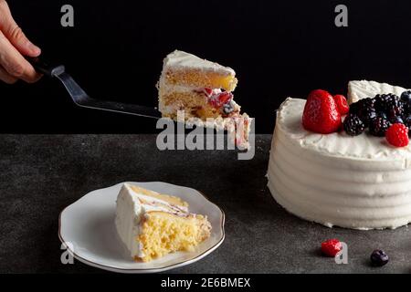 A round cake with creamy white icing and assortment of berries used as decorative toppings. Served On dark gray black background. A slice is on a plat Stock Photo