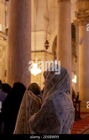 An Arabic Muslim woman wearing floral traditional head wrap is praying by raising her hands above inside a mosque in Syria. In the background there ar Stock Photo