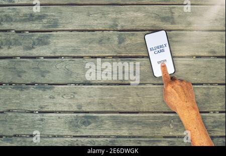 Enter Elderly Care app with senior hand touching the mobile screen on wooden table Stock Photo