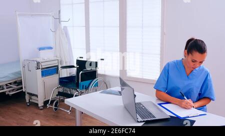 Woman therapist sitting at workplace looking at laptop reading news and taking notes on clipboard. Medical physician in medicine uniform writing list of consulted, diagnosed patients, making research. Stock Photo