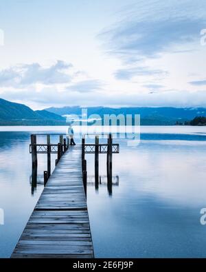Lonely woman standing at the end of Lake Te Anau jetty, looking at the Murichison mountains. Vertical format. Stock Photo