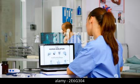 Doctor assistant analyzing digital human skeleton on laptop, taking notes on clipboard. Radiologist nurse in medicine uniform looking at digital radiography, bones examination, diagnosis Stock Photo