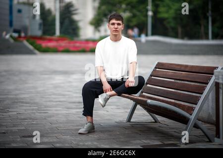 White casual t-shirt mockup on a guy sitting against a blurred park background, on a bench, front view, for design presentation, logo. Blank textured Stock Photo