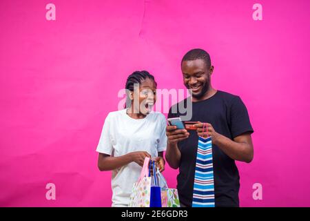 young black beautiful lady holding some gift bags with a beautiful smile and a man making online payment with his card on his smartphone Stock Photo