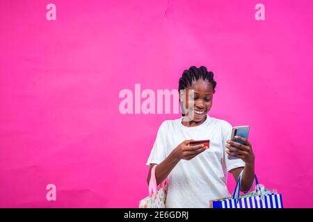 young black beautiful lady holding some gift bags with a beautiful smile and making online payment with her card on her smartphone Stock Photo