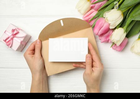 Female hands hold envelope with blank card on white wooden background with tulips and gift box Stock Photo