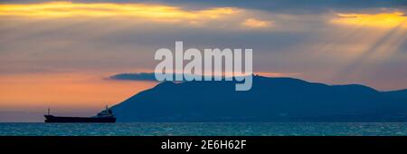 Beautiful sunset amongst the clouds. The sun rays are simply beautiful. City, mountains and ship in sea. Stock Photo