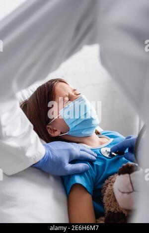 diseased girl in medical mask lying with closed eyes near doctor examining her with stethoscope, blurred foreground Stock Photo