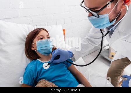 pediatrician in medical mask examining sick girl with stethoscope in hospital Stock Photo