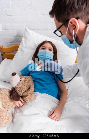 sick girl in medical mask lying in bed with teddy bear while doctor examining girl with stethoscope, blurred foreground Stock Photo