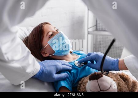 diseased girl in medical mask lying in bed with teddy bear while pediatrician examining her with stethoscope, blurred foreground Stock Photo