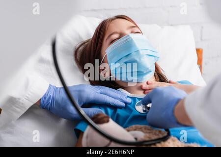 doctor examining girl, lying in medical mask with closed eyes, with stethoscope, blurred foreground Stock Photo