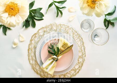 Effortless golden birthday dinner table decor. Pale yellow peony flowers and Late Spring, Summer flat lay. White dinner table, white, gold utensils Stock Photo