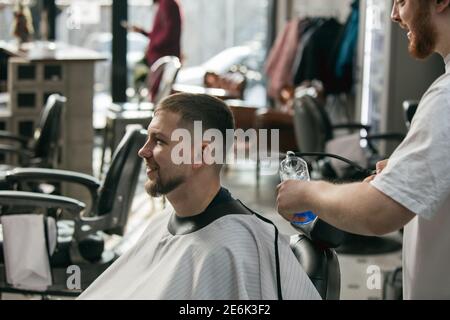 Master barber, stylist does the hairstyle to delighted smiling guy, young man. Professional occupation, male beauty concept. Cares of hair, mustache, beard of client. Soft colors and focus, vintage. Stock Photo