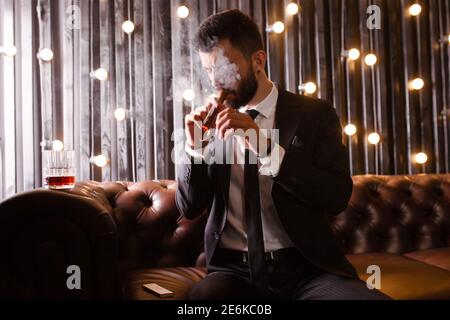 Portrait of a brutal beard man with tattoo smoking cigar in hot elegant suit  drinking whiskey alcohol in loft apartment . Couple love story concept Stock Photo