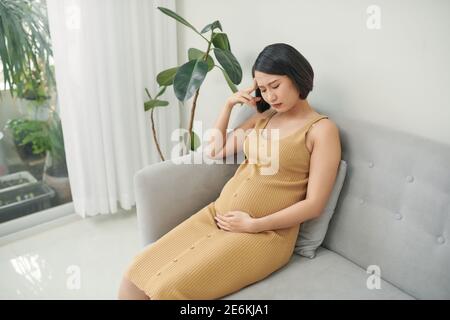 Young pregnant woman with headache sitting on sofa. Pregnancy symptoms, expectation, parenthood concept, copy space Stock Photo