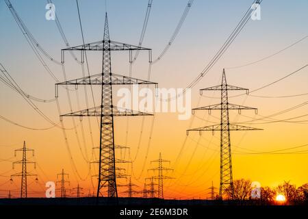 Electricity pylons and electric power transmission lines against vibrant orange sky at sunset. High Voltage towers provide power supply over a long di Stock Photo
