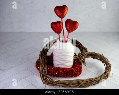 Wooden heart wreath, red glittery wreath and a white vase filled with red glitter heart sticks sitting on a herringbone tile counter with a plaster ba Stock Photo