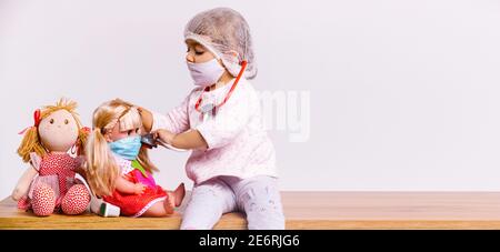 Banner,long format. A cute girl plays a doctor in a white coat, in a mask and with a stethoscope treats dolls. White background and side advertising Stock Photo
