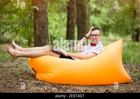 Scared student in glasses with laptop computer lying on inflatable mattress outdoor in summer park. Education, business, fail and technology concept Stock Photo